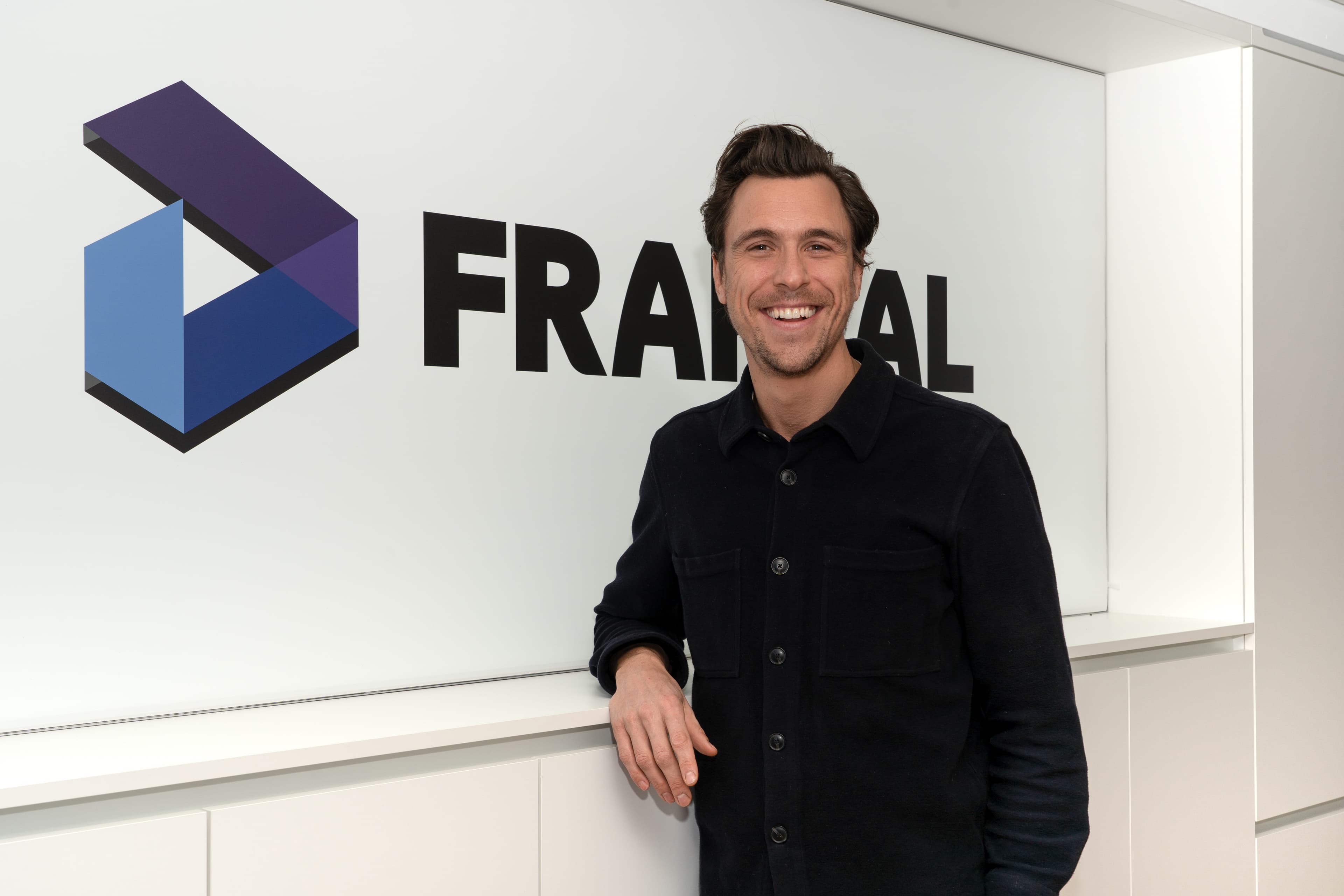 Kilian v. Dallwitz, the Press Spokesperson for Fraktal Development, presents a cheerful demeanor with a broad smile. He's dressed in a casual yet smart dark shirt, lending an approachable yet professional image. The 'FRAKTAL' logo looms prominently in the background, signifying the brand's strong presence.