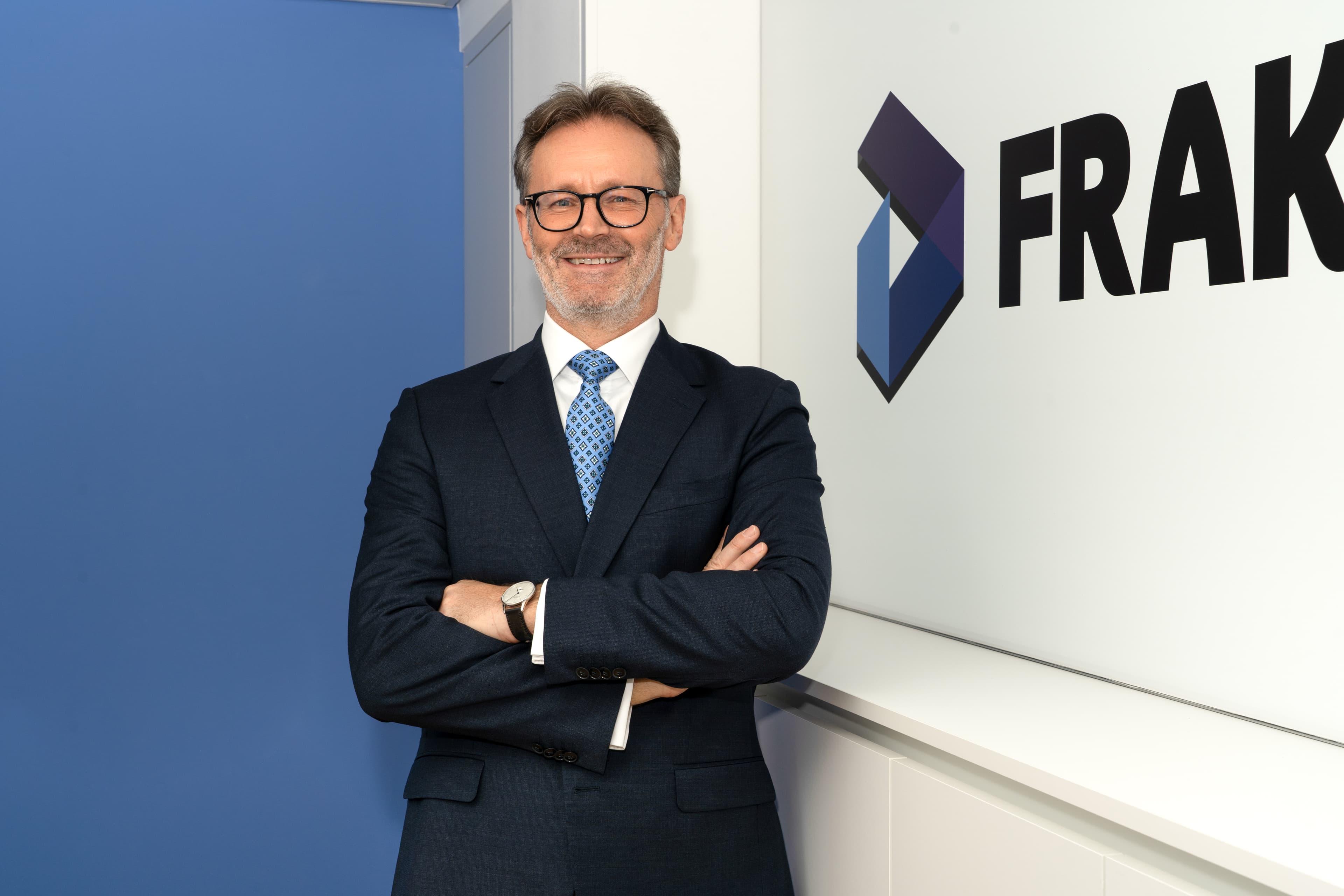 Portrait of Anthony Pink from Fraktal Development, poised confidently with arms folded in front of a panel displaying the FRAKTAL logo. He is dressed in a professional navy blue suit, patterned blue tie, and glasses, offering a friendly smile in a well-lit office setting that reflects the company branding colors.