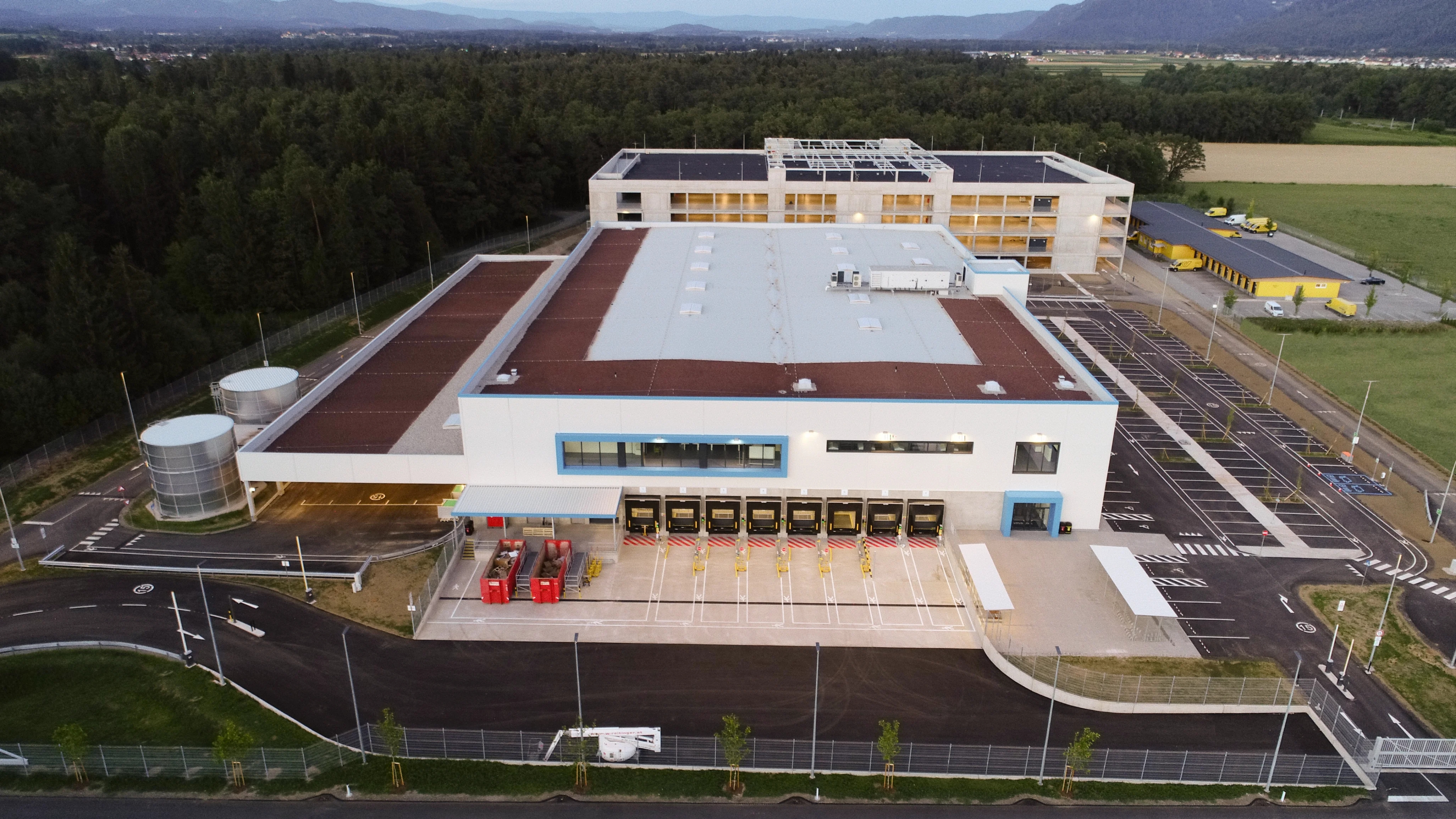 Aerial view of Fraktal Development's project featuring a sprawling industrial complex at dusk, with multiple docking stations for yellow trucks, a spacious parking area, and surrounding greenery, against a scenic backdrop of distant mountains and a fading evening sky.